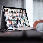 Auf einem Schreibtisch steht ein 2-in-1-Laptop auf dem ein digitales Meeting zu sehen ist. Davor sitzt eine Person, von der nur die Hände zu sehen sind.