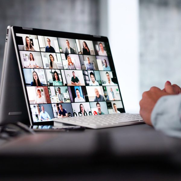 Auf einem Schreibtisch steht ein 2-in-1-Laptop auf dem ein digitales Meeting zu sehen ist. Davor sitzt eine Person, von der nur die Hände zu sehen sind.