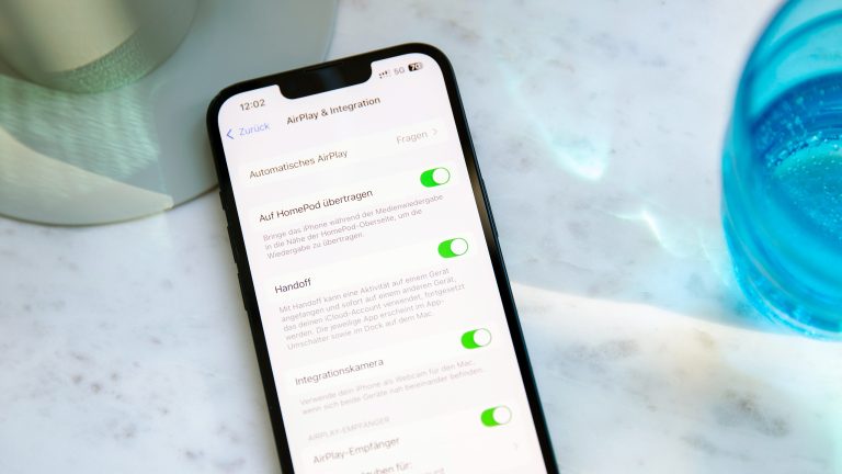 AirPlay, So funktioniert das drahtlose Streaming von Apple Ein iPhone liegt neben einem Wasserglas auf einer Marmorplatte. Darauf sind die AirPlay- und Integrations-Einstellungen zu sehen.