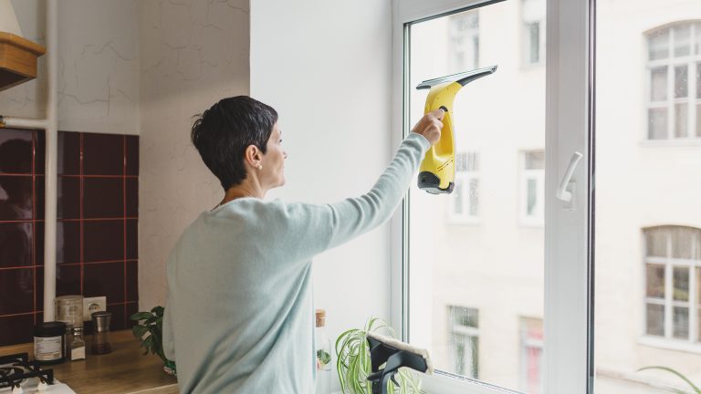 Eine Person steht in einer Küche vor dem Fenster und zieht mit einem Fenstersauger die Fensterscheibe von innen ab.