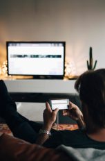Eine Person sitzt auf einer Couch in einem Wohnzimmer und hält ihr Smartphone quer in den Händen. Auf einem Fernseher vor ihr sieht man den gleichen Inhalt, wie auf dem Smartphone.
