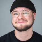 Eine Person mit schwarzer Mütze, Bart und goldener Brille.