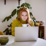 Eine Person sitzt am Küchentisch vor ihrem Laptop. In der linken Hand hält die Person eine Tasse.