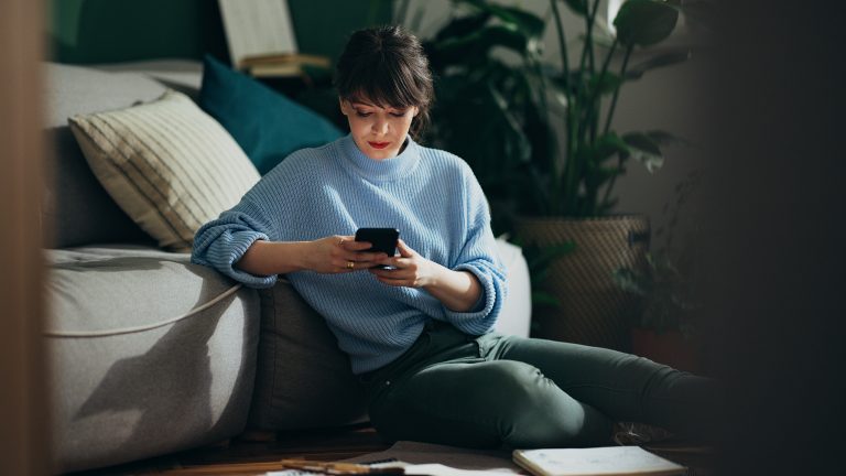 eSIM vs. Klassische SIM, Unterschiede, Vor- und Nachteile Eine Person sitzt im Halbdunkel auf dem Boden vor dem Sofa und hält ein Smartphone in den Händen.