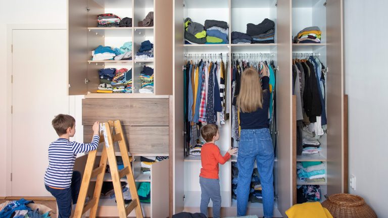 Eine Person steht mit zwei Kindern vor einem deckenhohen Kleiderschrank mit vielen Fächern und geöffneten Türen.