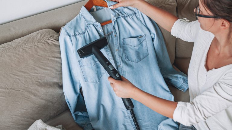 Eine Person behandelt mit einem Steamer ein Hemd, das sie gegen das Kissen auf einem Sofa lehnt.