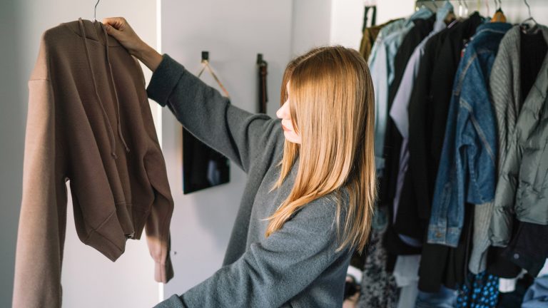 Eine Person steht vor einer Kleiderstange mit Jacken, hält einen Kleiderbügel mit einem Pullover vor sich und begutachtet diesen.