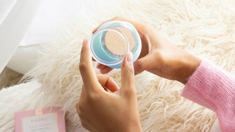 Home Spa, Diese Gadgets sorgen für Wellness zu Hause Die Hände einer Person öffnen das FOREO UFO über einer weißen Kuscheldecke. Daneben liegt die Verpackung der Maske „Glow Addict“ von FOREO.