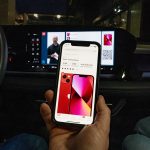 Eine Person sitzt in einem Audi Q6 e-tron und hält ein Smartphone in der Hand. Darauf ist eine Produktdetailseite aus dem OTTO-Shop zusehen und im Hintergrund läuft auf dem integrierten Display ein Live-Shopping-Stream.