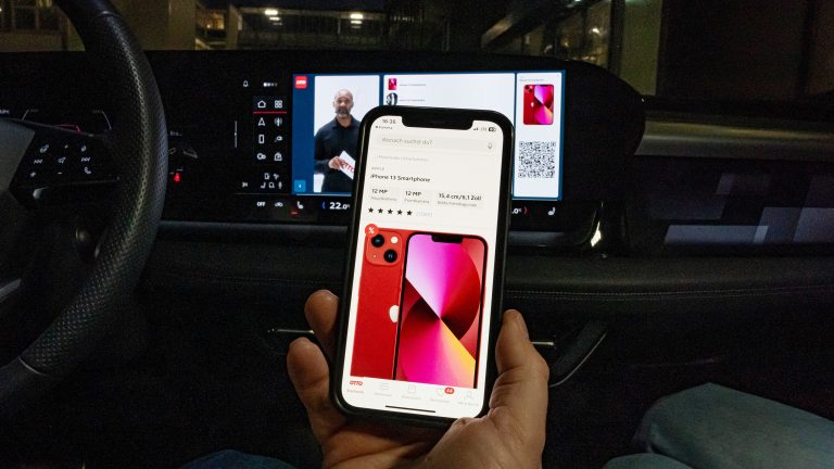 Eine Person sitzt in einem Audi Q6 e-tron und hält ein Smartphone in der Hand. Darauf ist eine Produktdetailseite aus dem OTTO-Shop zusehen und im Hintergrund läuft auf dem integrierten Display ein Live-Shopping-Stream.
