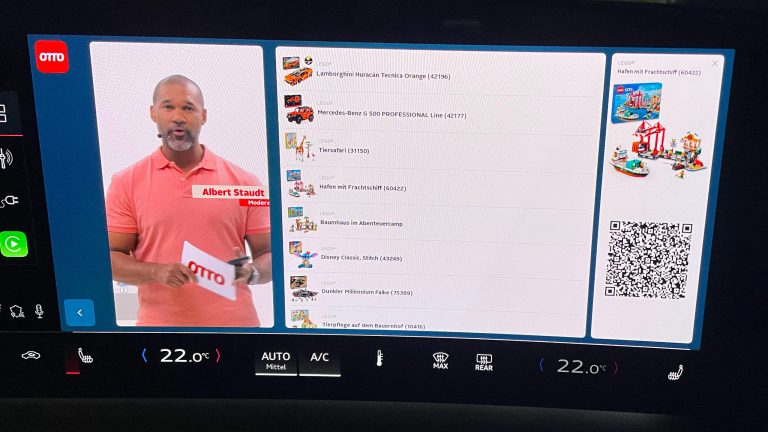 Erlebe OTTO Live Shopping im Audi Q6 e-tron! Eine Nahaufnahme eines integrierten Displays in einem Auto. Darauf ist ein Live Shopping Stream von OTTO zu sehen: links ein Moderator, mittig eine Auflistung von Produkten und rechts ein einzelnes Produkt mit einem QR Code.