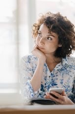 Struktur im Alltag, Tools für mehr Produktivität Eine Person sitzt vor einem großen Fenster an einem Tisch und hält ein Smartphone in der Hand.