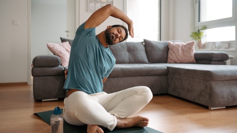 Struktur im Alltag, Tools für mehr Produktivität Eine Person sitzt im Schneidersitz vor einem Sofa auf einer Yogamatte und dehnt sich.