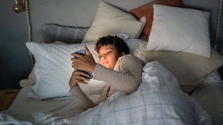 Struktur im Alltag, Tools für mehr Produktivität Eine Person liegt in einem Bett mit vielen Kissen, hält ihr Smartphone mit beiden Händen vors Gesicht und wird davon erleuchtet.