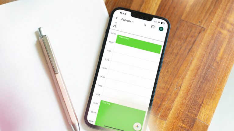 Struktur im Alltag, Tools für mehr Produktivität Ein Smartphone liegt halb auf einem Notizbuch, halb auf einem Holztisch. Darauf ist eine Tagesübersicht eines Kalenders mit zwei Terminen darin zu sehen. Neben dem Handy liegt ein Stift.