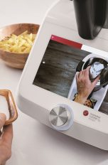 Die Hände einer Person halten eine kleine Schüssel und einen kleinen Löffel, der in die Monsieur Cuisine smart-Küchenmaschine gegeben werden soll, die auf der Küchentheke steht.