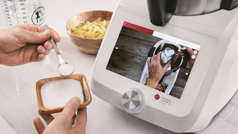 Thermomix-Alternative, Modelle im Vergleich Die Hände einer Person halten eine kleine Schüssel und einen kleinen Löffel, der in die Monsieur Cuisine smart-Küchenmaschine gegeben werden soll, die auf der Küchentheke steht.