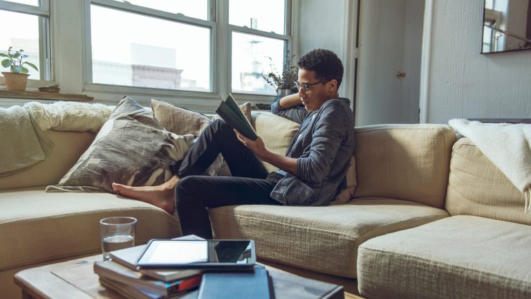 Kindle Unlimited, So kündigst du das E-Book-Abo Eine Person sitzt auf einer Couch vor einem breiten Fenster und hält ein Buch in der Hand. Auf dem Couchtisch davor stapeln sich zahlreiche Bücher, auf denen ein Tablet liegt.