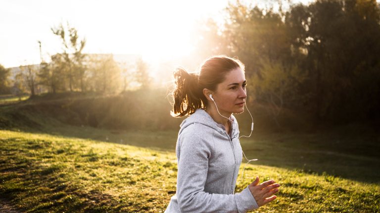 Podcast Apps, Diese Apps gibt es Eine Person joggt an einer Wiese entlang. Im Hintergrund ein paar Bäume und direkte Sonne.