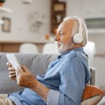 Eine ältere Person sitzt auf einem Sofa, schaut auf ihr Tablet und trägt dabei Kopfhörer.