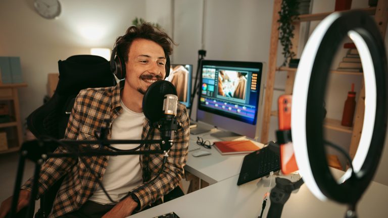 Eine Person sitzt an einem Schreibtisch mit einem professionellen Set-Up, um Content zu produzieren. Zu sehen ist unter anderem ein Ringlicht, ein Mikrofon und eine Smartphone-Halterung und ein großer Monitor.