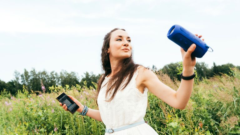 Eine Person steht auf einer Wiese. In einer Hand hält sie einen Bluetooth-Lautsprecher, in der anderen Hand ein Smartphone.