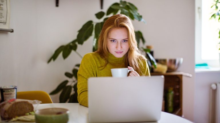 Wasserzeichen in Word einfügen und bearbeiten Eine Person sitzt an einem runden Tisch vor einem geöffneten Laptop. In der linken Hand hält sie eine Kaffeetasse.