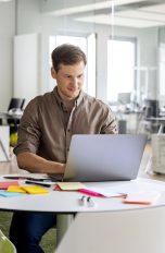 Eine Person sitzt in einem Großraumbüro an einem runden Tisch, auf dem ein geöffnetes Notebook steht.