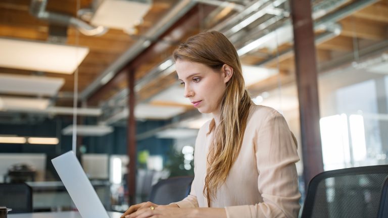 Zeitstrahl in PowerPoint einfügen und bearbeiten, Schritt für Schritt Seitenansicht einer Person, die in einem Büro an einem Schreibtisch vor einem geöffneten Laptop sitzt.
