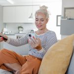 Eine Person sitzt auf der Couch, schaut auf ihr Smartphone in der einen Hand und richtet mit der anderen eine Fernbedienung nach vorn.
