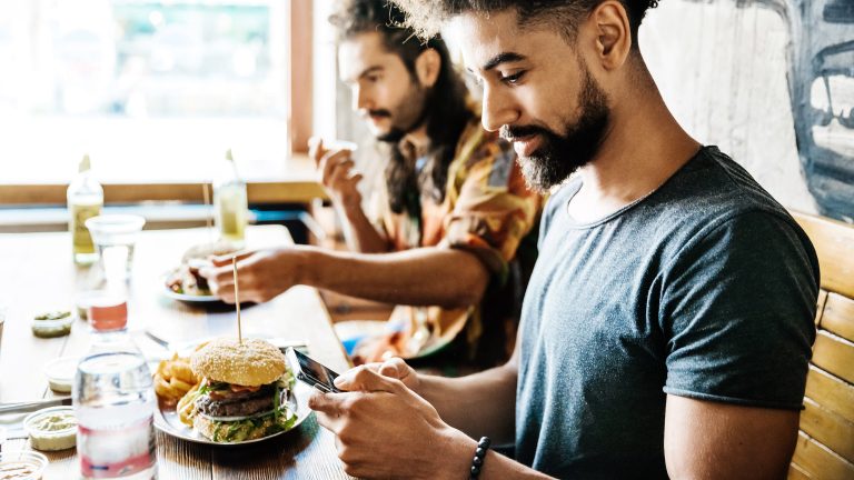 Apple Pay, Bezahlen mit iPhone, iPad und Apple Watch Zwei Personen sitzen am Tisch und Essen. Eine Person bedient dabei ihr Smartphone.