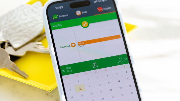 Auf einem Smartphone ist die App „Ausgaben Manager-Tracker“ geöffnet. Im Hintergrund ist eine gelbe Ablage mit einem Schlüsselbund zu sehen.