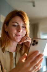Eine Person hält ein Smartphone in ihrer Hand, in welches sie hineinblickt. Im Hintergrund ist eine Kaffeetasse sowie ein Teil einer Küche zu erahnen.