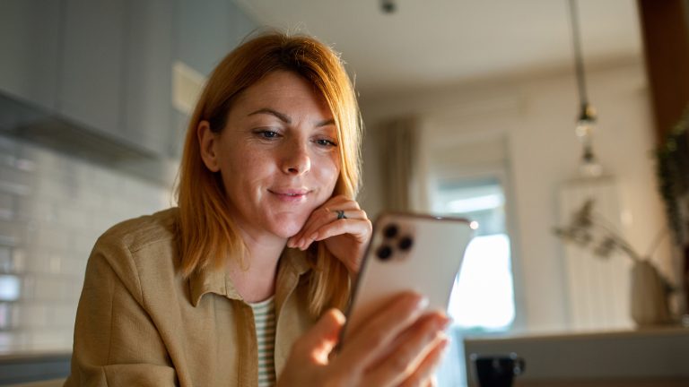 Banking-Apps, 5 Anwendungen für iOS & Android für easy Online-Banking Eine Person hält ein Smartphone in ihrer Hand, in welches sie hineinblickt. Im Hintergrund ist eine Kaffeetasse sowie ein Teil einer Küche zu erahnen.