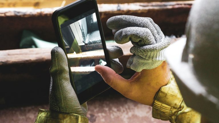 Das sollte dein Baustellenhandy können Nahaufnahme von Händen, die in halb abgestreiften Arbeitshandschuhen stecken um ein Smartphone zu bedienen.