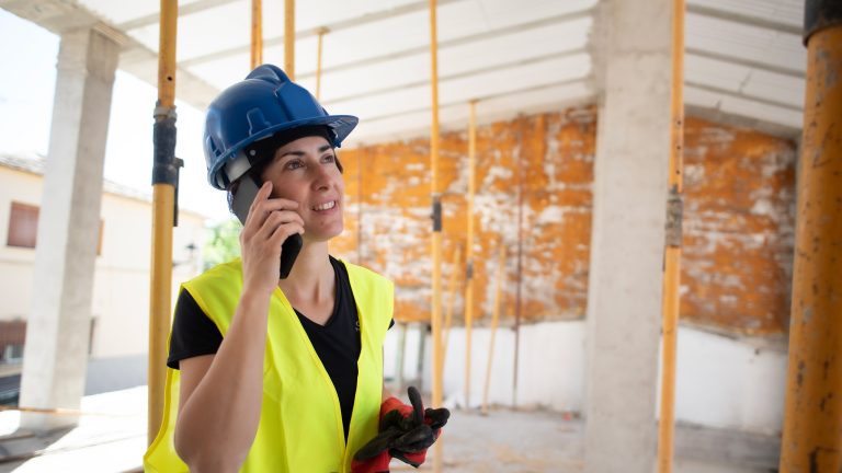 Das sollte dein Baustellenhandy können Eine Person steht mit einer Sicherheitsweste und einem Schutzhelm bekleidet auf einer Baustelle und telefoniert mit ihrem Smartphone.