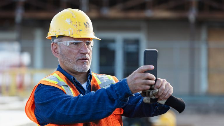 Das sollte dein Baustellenhandy können Eine Person mit einer Sicherheitsweste und einem Schutzhelm, hält ein Smartphone in den Händen.