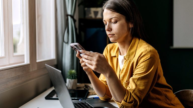 Eine Person sitzt an einem Schreibtisch, auf dem ein ge&ouml;ffneter Laptop steht. Sie schaut in ein Smartphone, das sie in beiden H&auml;nden h&auml;lt.