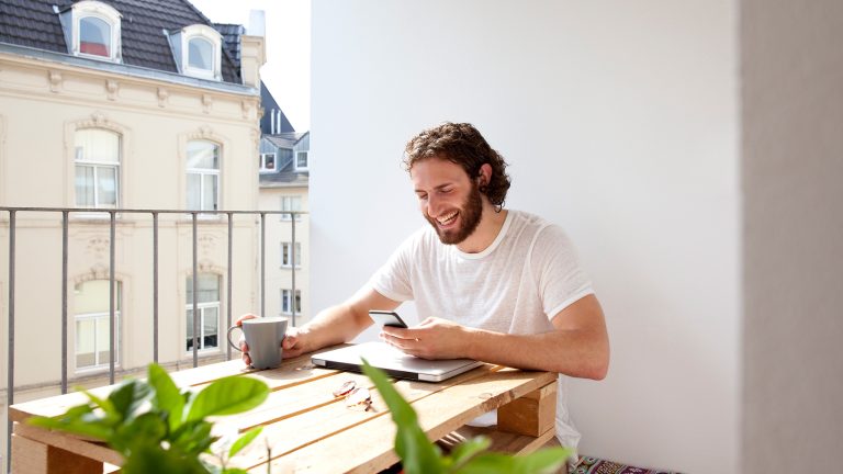 Facebook-Videos downloaden, So klappt’s Eine Person sitzt vor einem zugeklappten Laptop und hat ein Smartphone in der Hand.