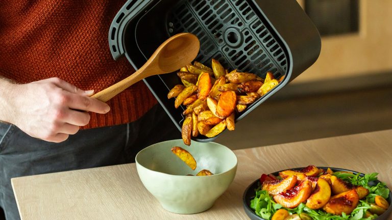 Die Hand einer Person hält einen Holzkochlöffel, um Kartoffelwedges aus dem Frittierkorb eines Airfryers in eine Schüssel zu geben. Daneben steht ein Salat mit Pfirsichvierteln.