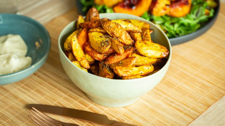 Frittieren ohne Fett, Funktionen einer Heißluftfritteuse Auf einem Tisch stehen drei mittelgroße Schüsseln mit Essen. Im Fokus stehen die Kartoffel-Wedges. Daneben liegt goldfarbenes Besteck.