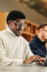Eine Person sitzt neben einer anderen Person an einem Tisch und tippt auf einem Laptop.