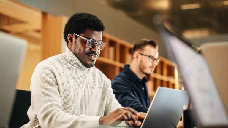 Eine Person sitzt neben einer anderen Person an einem Tisch und tippt auf einem Laptop.
