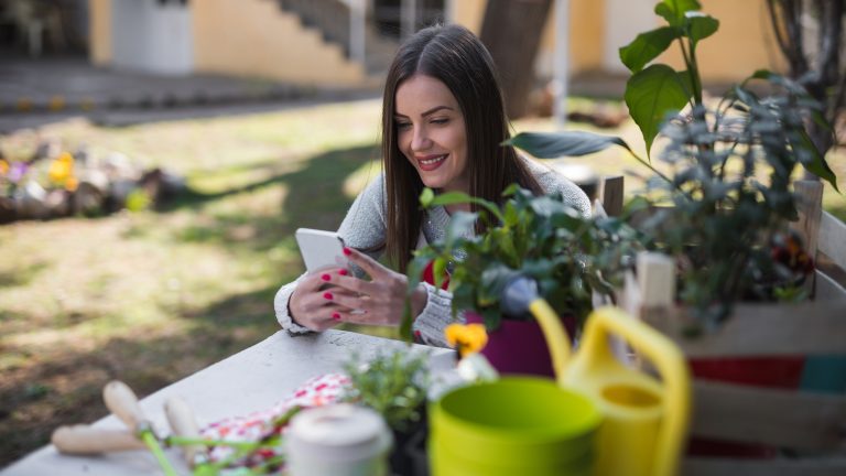 So machst du deinen Garten im Frühling fit Eine Person sitzt im Garten an einem Tisch, auf dem verschiedene Werkzeuge zur Gartenarbeit liegen. In den Händen hält sie ein Smartphone.