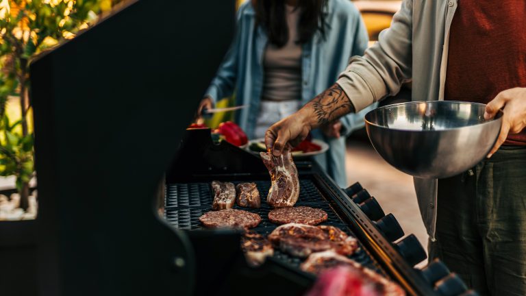 So kannst du deinen Gasgrill reinigen Seitenansicht eines Gasgrills auf dem bereits Steaks und Pattys liegen. Eine Person, die eine Schüssel in der linken Hand hält, ist gerade dabei mit der rechten Hand ein weiteres Steak dazu zu legen.