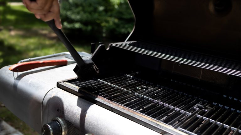 So kannst du deinen Gasgrill reinigen Es ist die Hand einer Person zu sehen, die mit einer Eisenbürste grobe Reste vom Grillrost entfernt.