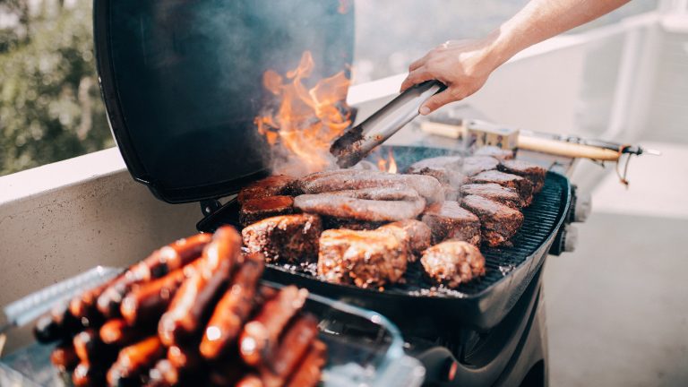 Auf dem Grillrost liegen zahlreiche Fleischstücke. Flammen bersten durch den Rost, darüber eine Hand mit einer Zange, um das Fleisch zu wenden.