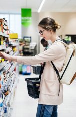 Eine Person steht vor einem Supermarktregal, aus dem sie gerade etwas herausnehmen möchte. In der anderen Hand hält sie einen Einkaufskorb.