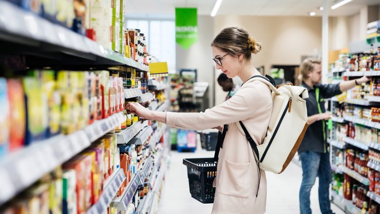 Eine Person steht vor einem Supermarktregal, aus dem sie gerade etwas herausnehmen möchte. In der anderen Hand hält sie einen Einkaufskorb.