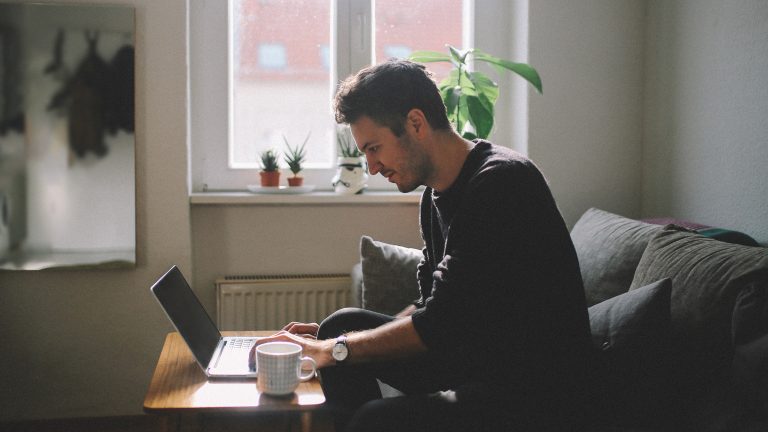 Laptop mit Fernseher verbinden, So geht‘s mit und ohne Kabel Eine Person sitzt auf einem Sofa in einem Wohnzimmer und tippt auf einem Laptop, der vor ihr auf einem kleinen Tisch steht.